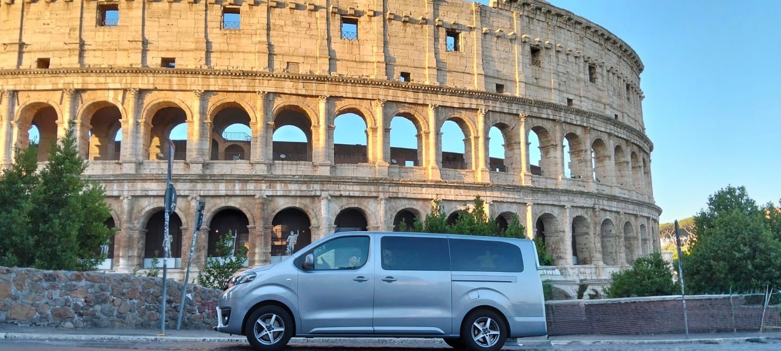 Srebrny van na tle Koloseum w Rzymie. Auto zaparkowane przy ulicy. Widok na starożytny amfiteatr w słoneczny dzień. Turystyka i transport.