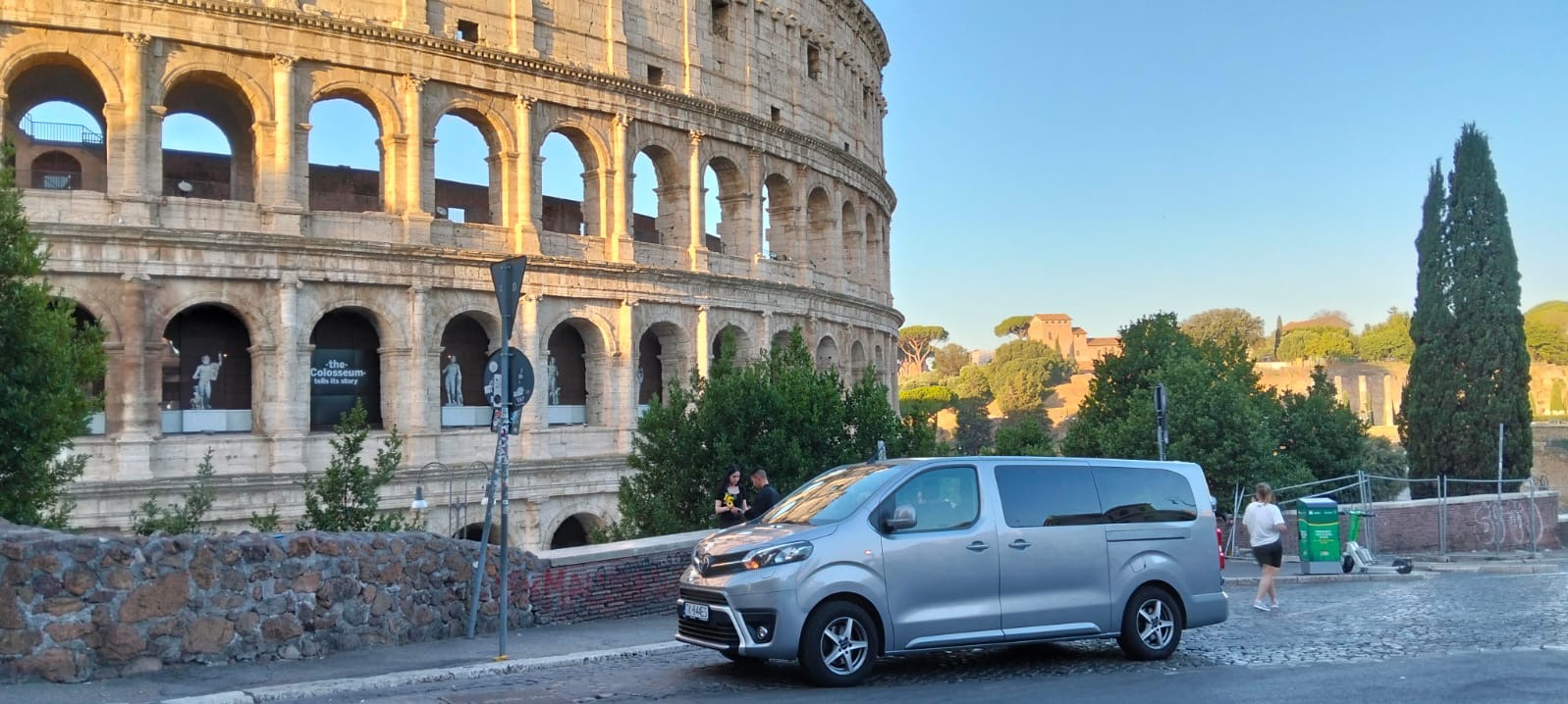Srebrny van na tle Koloseum w Rzymie, z zielonymi drzewami i fragmentem muru. Turystka przechodzi obok, a w tle widać zabudowania i niebo.