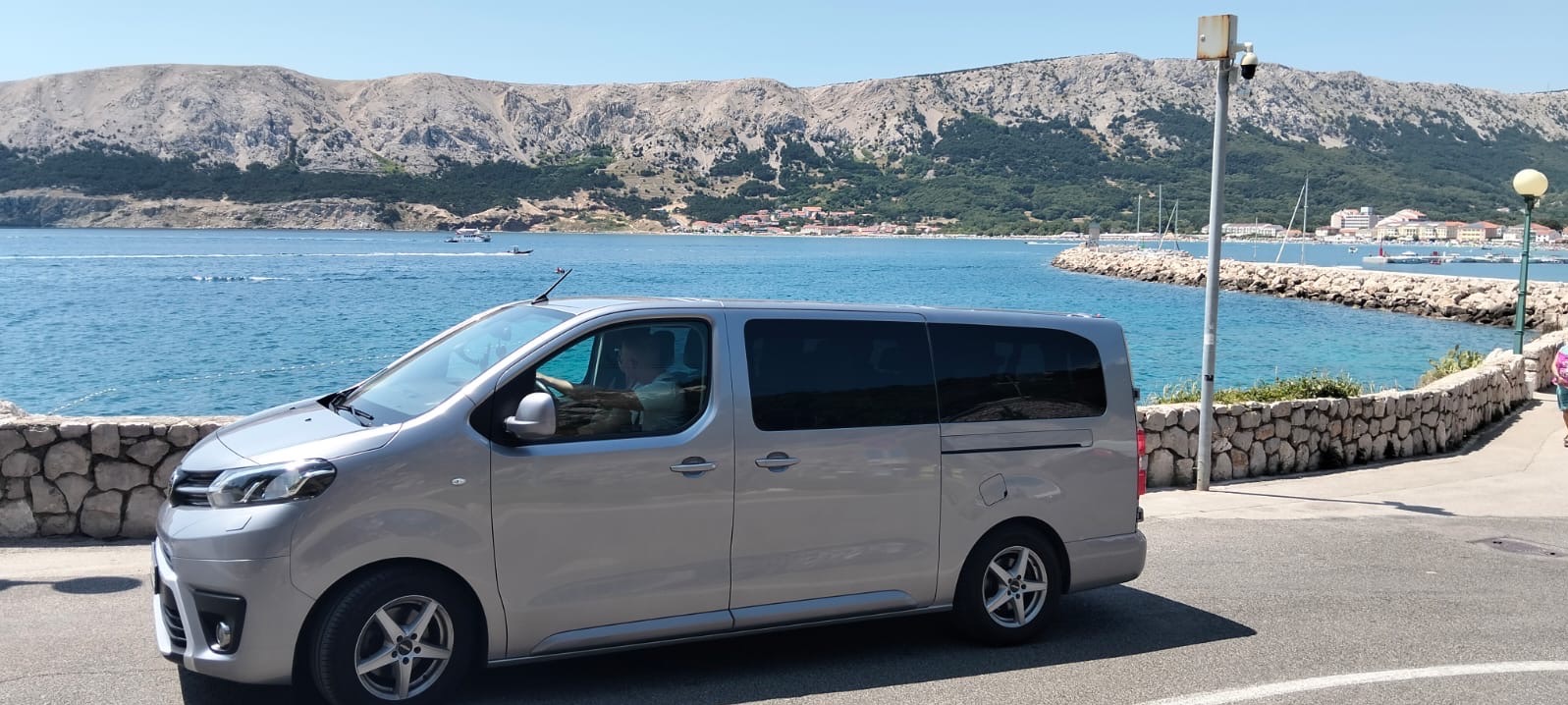 Srebrny van z kierowcą na tle malowniczego krajobrazu z morzem, górami i portem. Auto zaparkowane przy kamiennym murku, w tle słup z kamerą.