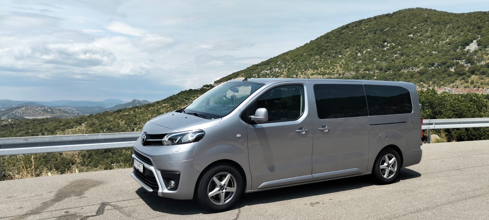 Srebrny van Toyota Proace Verso z przyciemnianymi szybami na tle górskiego krajobrazu. Auto zaparkowane na poboczu, widok z boku, lekko z góry. Jasne, naturalne światło.