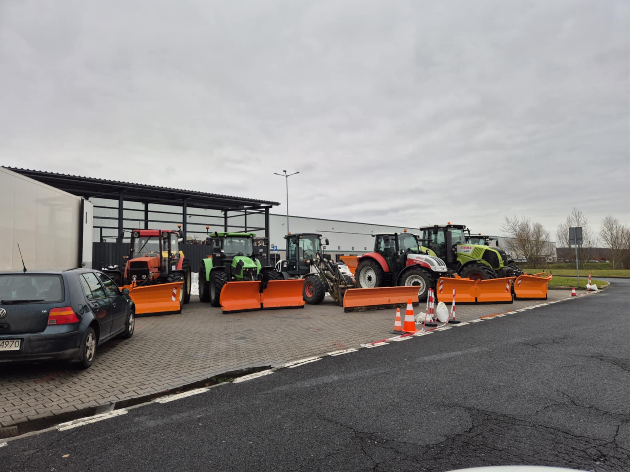 Flota traktorów z pomarańczowymi pługami do odśnieżania, zaparkowana na kostce brukowej obok asfaltowej drogi w pochmurny dzień. W tle budynek.