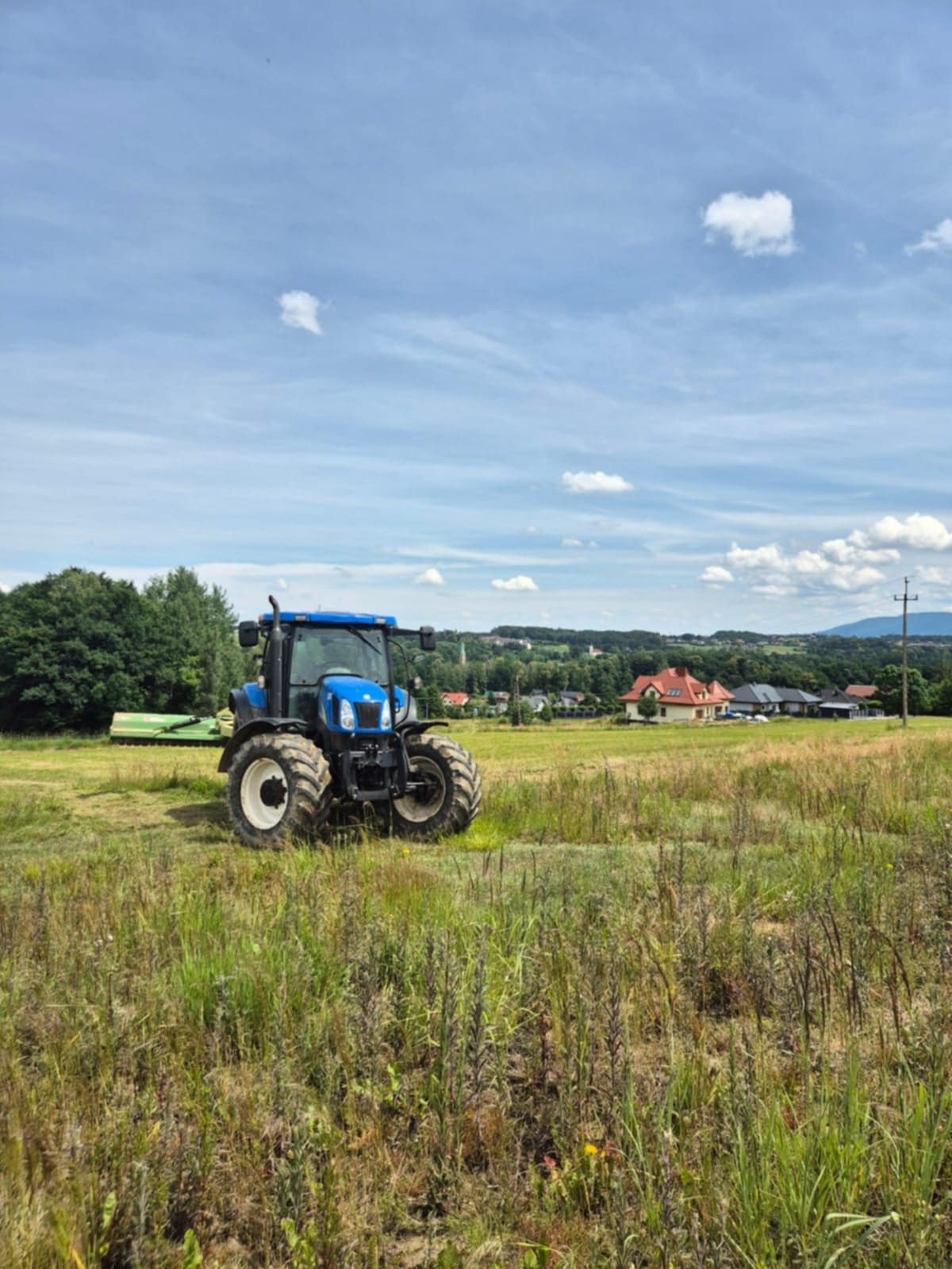 Niebieski traktor z kosiarką na łące, po koszeniu trawy, w tle zabudowania i błękitne niebo z chmurami. Widok na wieś.