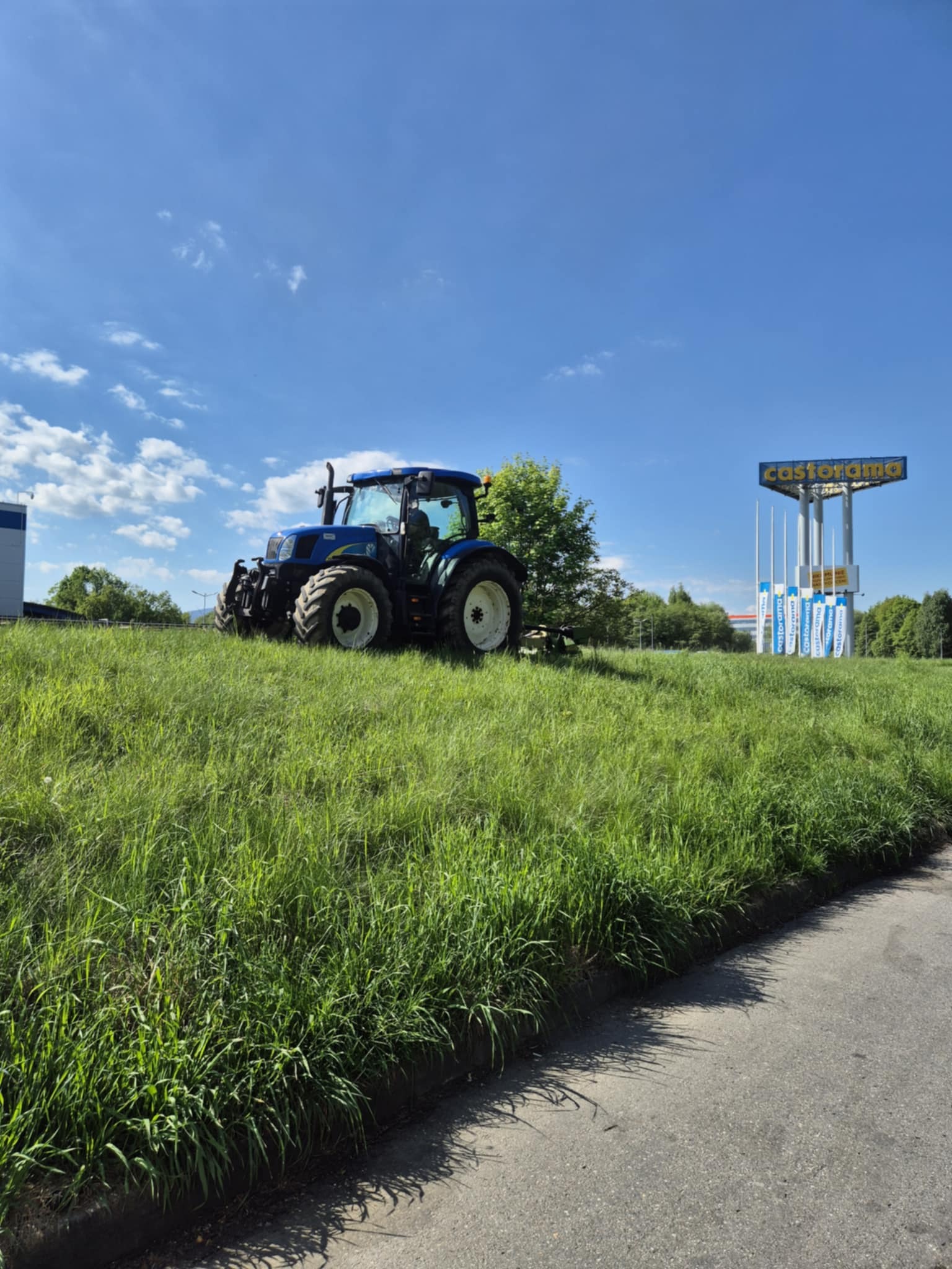 Niebieski traktor na wzgórzu z wysoką trawą, obok drogi i reklamy Castorama, w słoneczny dzień. Maszyna gotowa do koszenia trawy.
