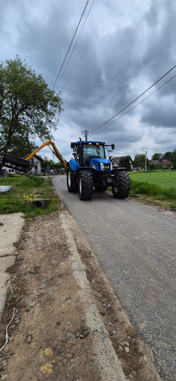Niebieski traktor z żółtym wysięgnikiem na asfaltowej drodze, obok trawnik i drzewa, pochmurne niebo. Sprzęt do odśnieżania dróg i placów.
