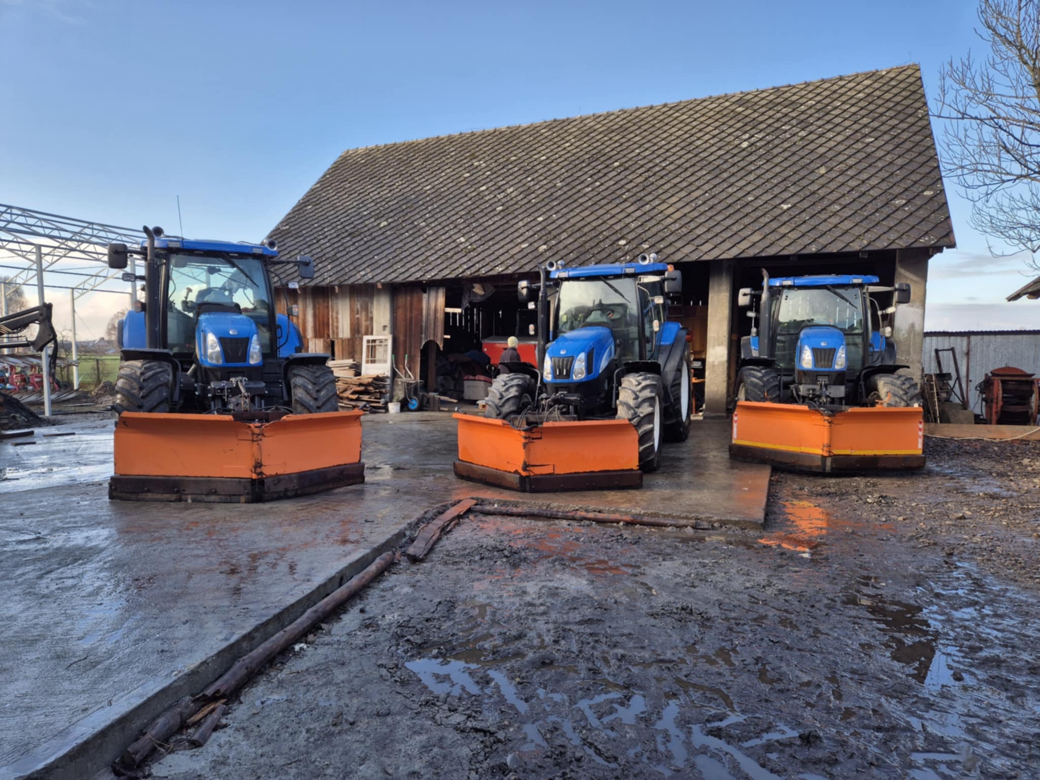 Trzy niebieskie traktory New Holland z pomarańczowymi pługami do odśnieżania stoją przed starym budynkiem gospodarczym w pochmurny dzień. Błotniste podłoże.