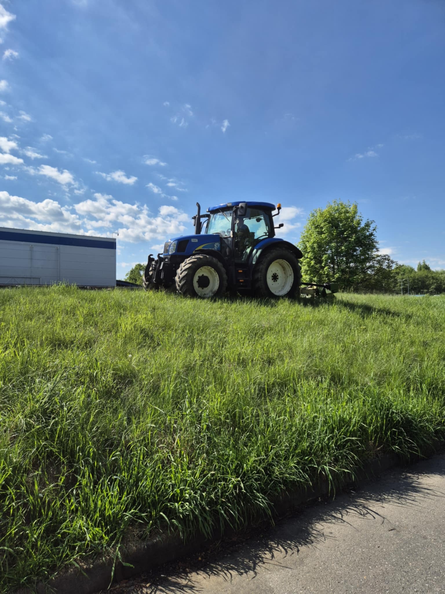 Niebieski traktor na tle błękitnego nieba z chmurami, stojący na wysokiej trawie, z fragmentem budynku w tle. Słoneczny dzień.