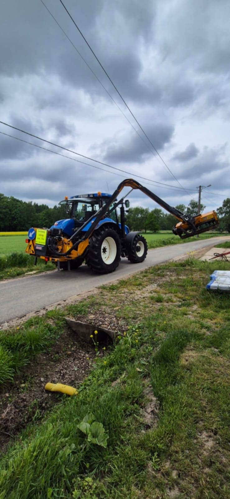 Niebieski traktor z wysięgnikiem i kosiarką boczną na asfalcie, obok zieleni. Kosiarka gotowa do użycia, w tle pola i pochmurne niebo.