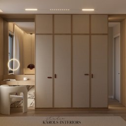 Karols Interiors - Neutralna kolorystyka szafy wnękowej z teksturowanymi frontami, zintegrowanej z toaletką z okrągłym lustrem podświetlanym LED, uzupełniona krzesłem boucle i dywanem w odcieniach beżu.