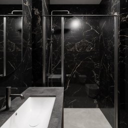 Studio Sawubona - Nowoczesna łazienka z czarnym marmurem, szklaną kabiną prysznicową i minimalistyczną umywalką. Metalowa bateria i subtelne oświetlenie podkreślają elegancję wnętrza.