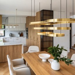 Studio Sawubona - Nowoczesna kuchnia z wyspą i drewnianymi akcentami. Złote lampy wiszące nad stołem. Parkiet w jodełkę. Minimalistyczny design z roślinami w wazonie.