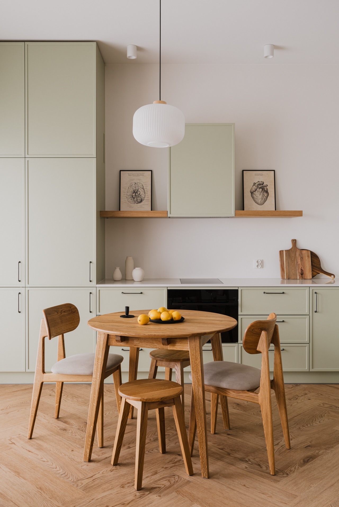 Jasna kuchnia z drewnianym stołem, krzesłami i stołkiem. Na stole czarne naczynie z cytrynami. Nad stołem wisi biała lampa. Na ścianie grafiki w czarnych ramkach.