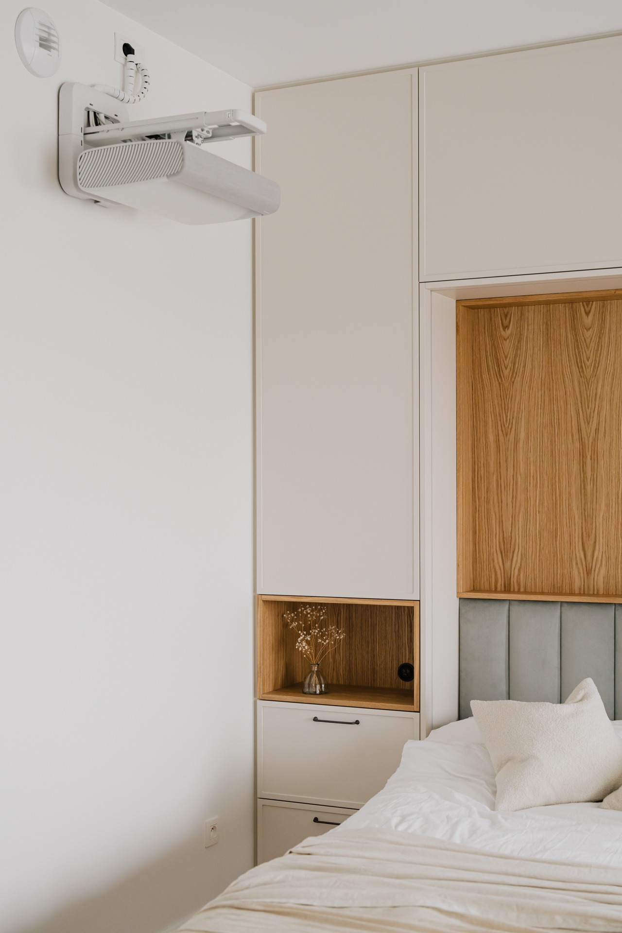 Nowoczesna sypialnia z ukrytym projektorem na ścianie, zabudowa meblowa z drewnianymi akcentami i subtelną dekoracją z suszonych kwiatów w szklanym wazonie.