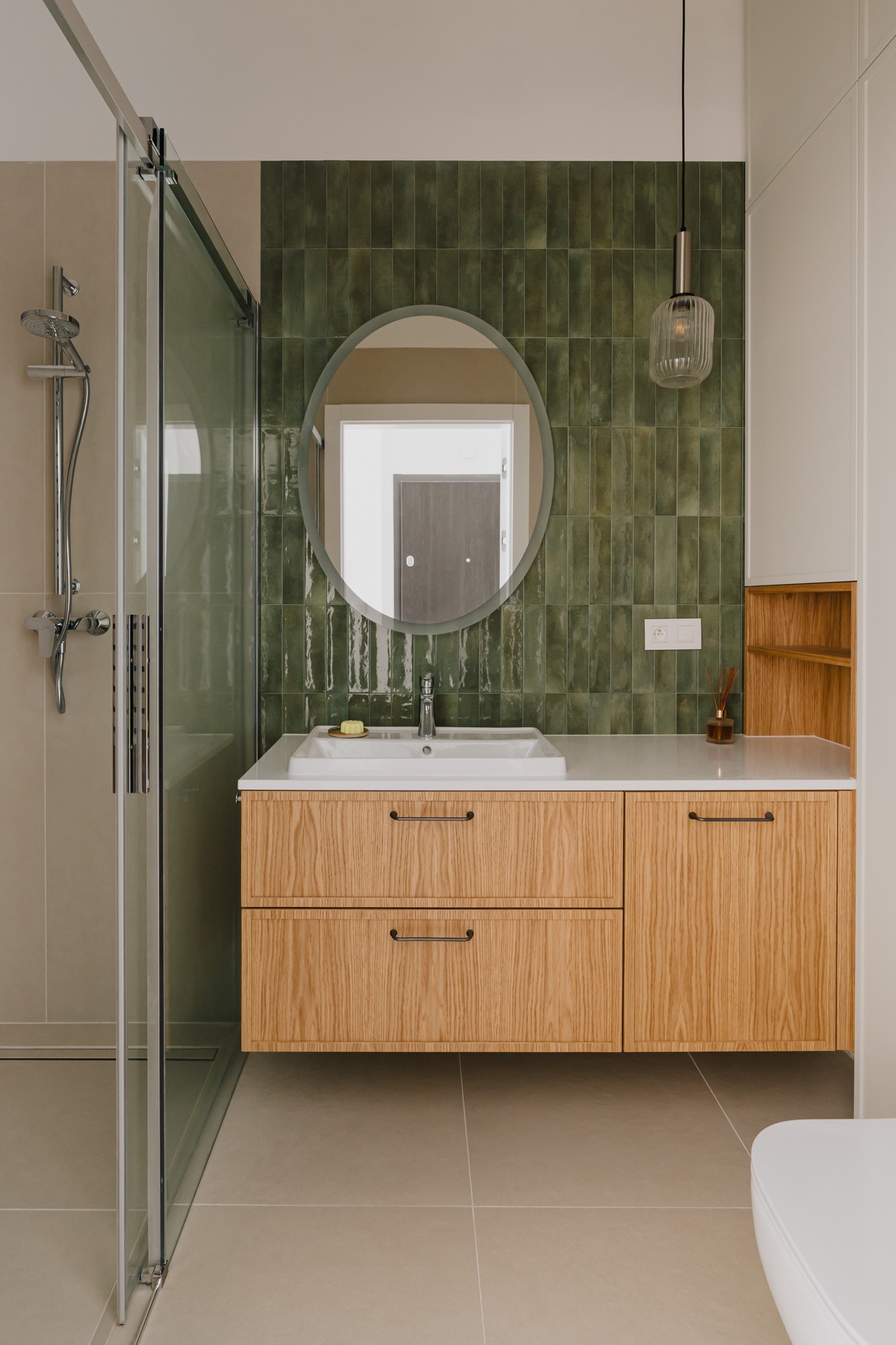 Nowoczesna łazienka z kabiną prysznicową ze szkła, drewnianą szafką pod umywalką z białym blatem, okrągłym lustrem i zieloną ścianą z płytek wertykalnych.