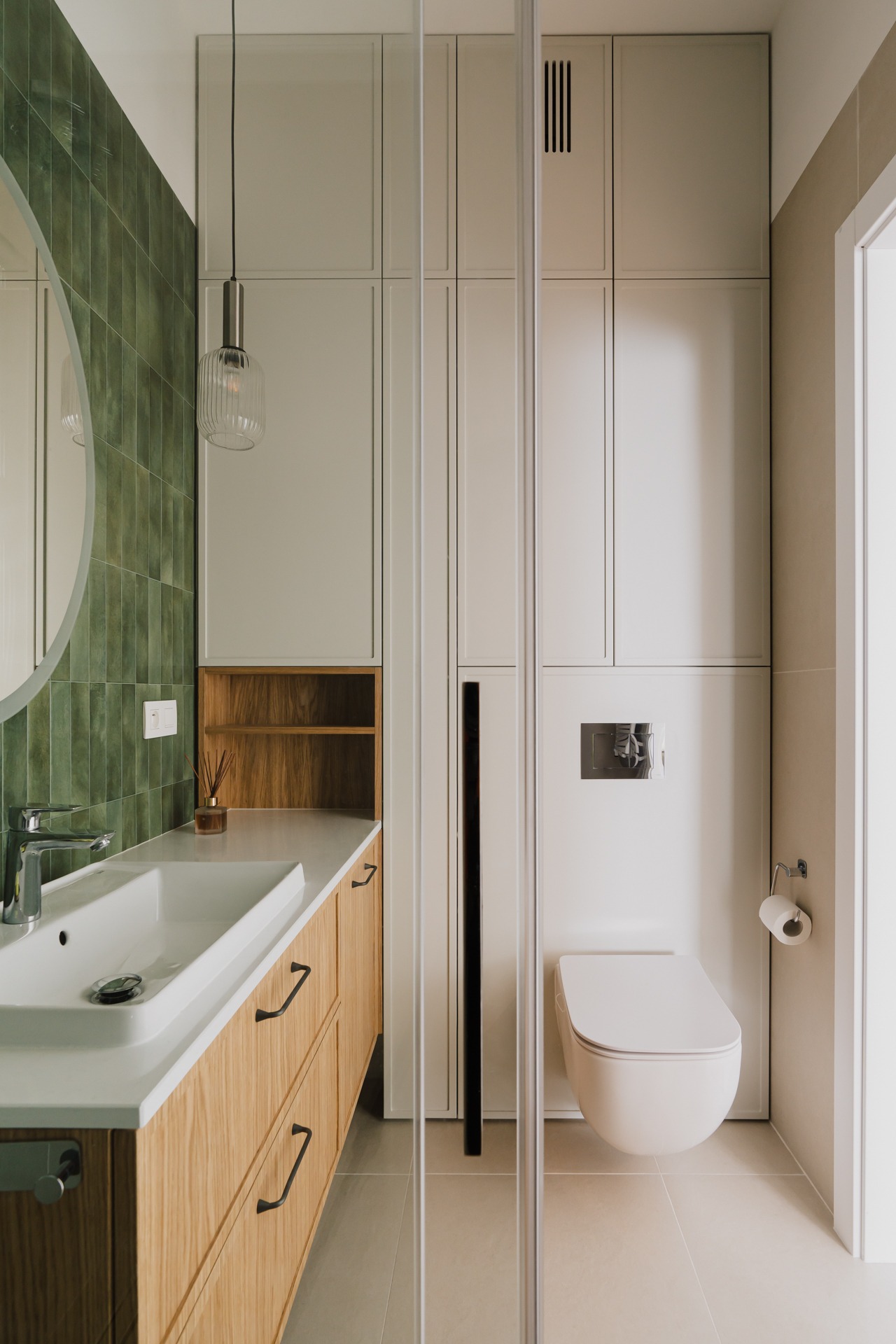 Nowoczesna łazienka z zielonymi płytkami na ścianie, drewnianą szafką pod umywalką, wiszącą toaletą i szklaną kabiną prysznicową, oświetlona wiszącą lampą ze szklanym kloszem.
