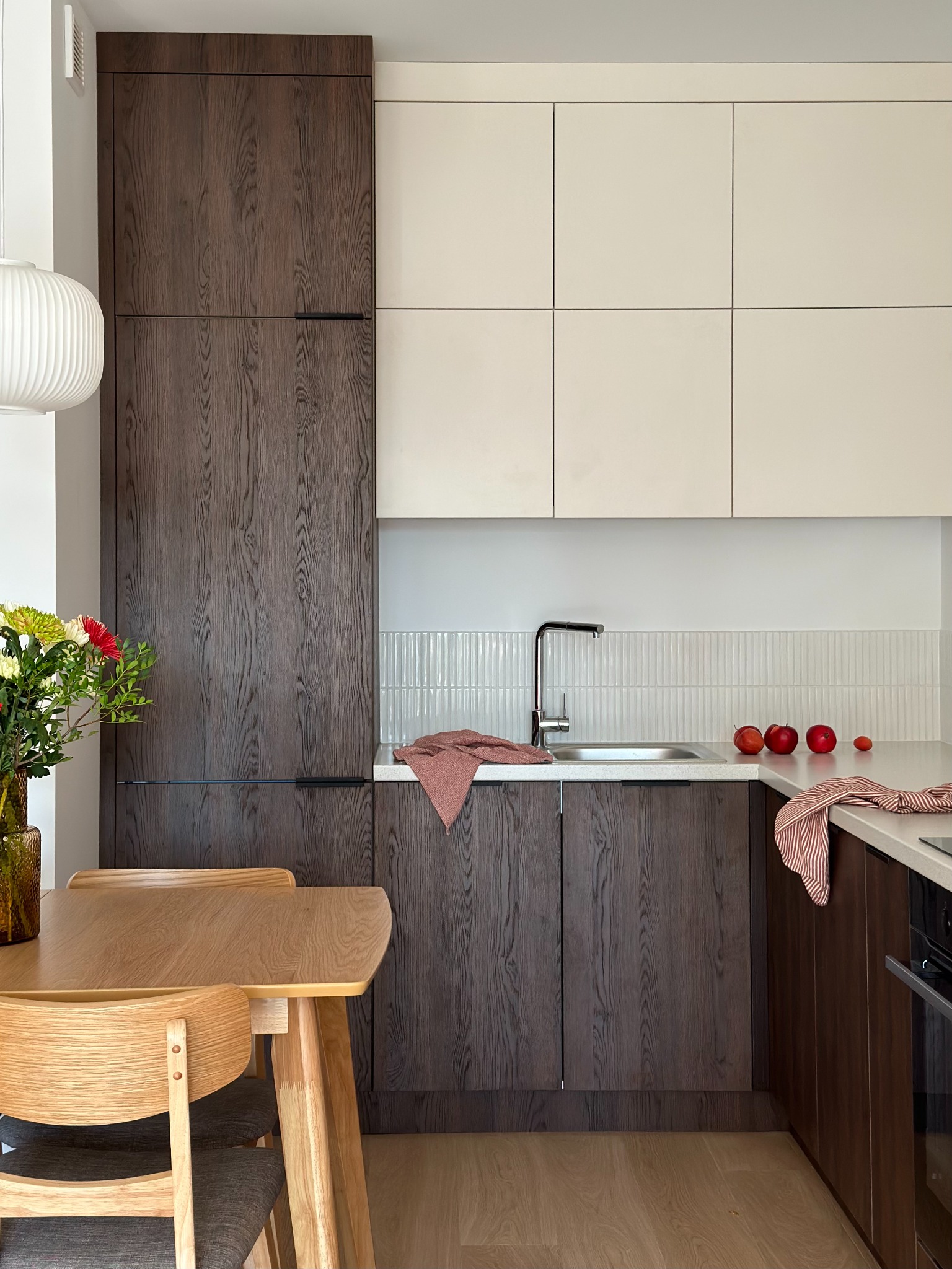 Nowoczesna kuchnia z drewnianymi frontami szafek w odcieniach brązu i beżu, minimalistyczny stół z krzesłami z jasnego drewna, owoce i kwiaty jako dekoracja, bateria kuchenna ze stali nierdzewnej.