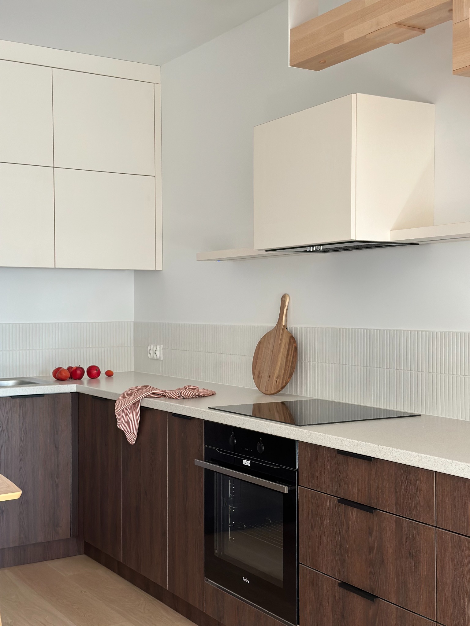 Minimalistyczna kuchnia z ciemnobrązowymi szafkami dolnymi i białymi górnymi, okrągła deska do krojenia zawieszona na ścianie z białych, pionowych płytek, na blacie leżą jabłka i ściereczka...