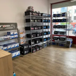 Wnętrze sklepu z akumulatorami samochodowymi, metalowe regały wypełnione różnymi modelami akumulatorów marek Exide i Grom Battery, widoczny laptop na biurku i logo Grom Battery z wizerunkiem orła.