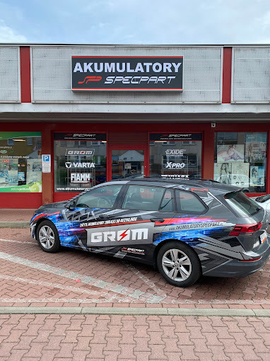 Samochód osobowy oklejony reklamą firmy 'Akumulatory Specpart' zaparkowany przed budynkiem firmy z szyldem 'Akumulatory Specpart' nad wejściem.