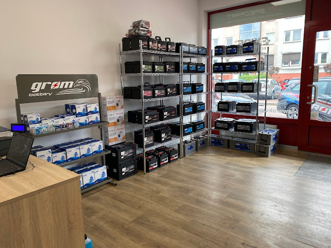 Wnętrze sklepu z akumulatorami samochodowymi, metalowe regały wypełnione różnymi modelami akumulatorów marek Exide i Grom Battery, widoczny laptop na biurku i logo Grom Battery z wizerunkiem orła.