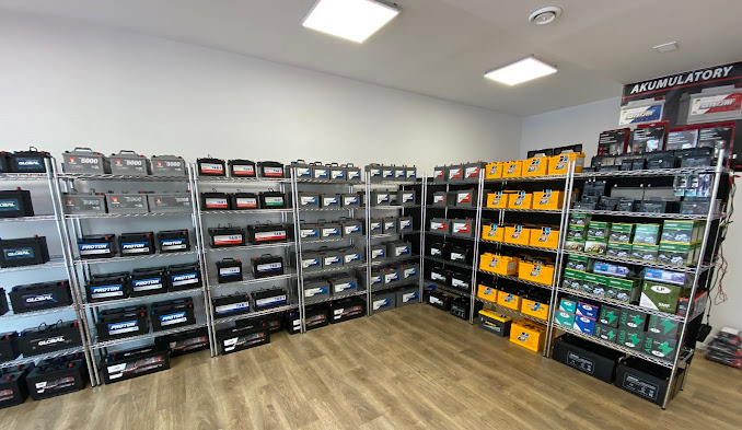 Sklep z akumulatorami samochodowymi, regały metalowe wypełnione różnymi markami i modelami, widoczne marki Global, Proton, TAB, Optima, ekspozycja w jasnym pomieszczeniu.