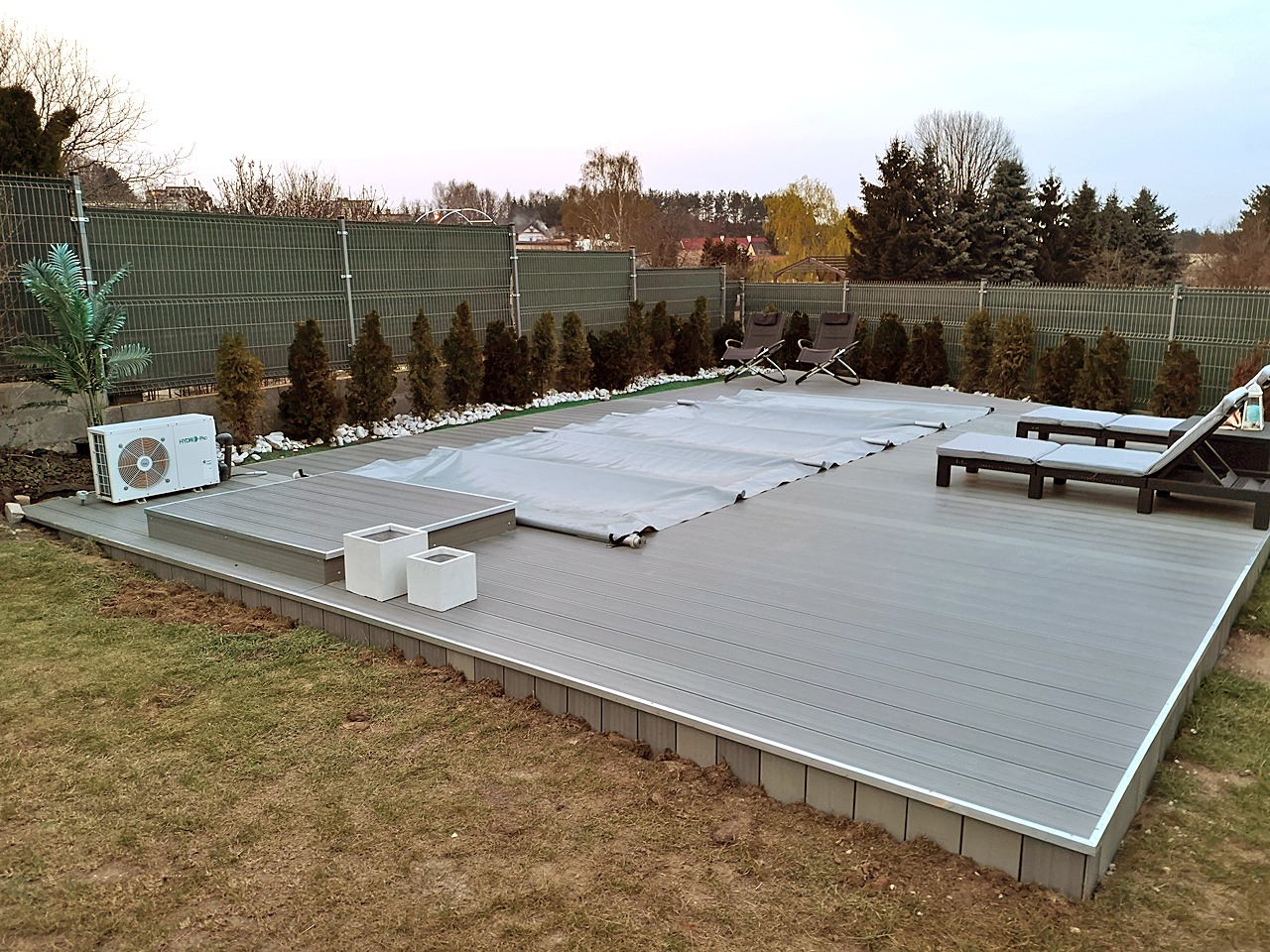 Szary taras kompozytowy otaczający basen przykryty plandeką, z leżakami i krzesłami ogrodowymi, widok z poziomu trawnika, w tle siatka ogrodzeniowa i drzewa.