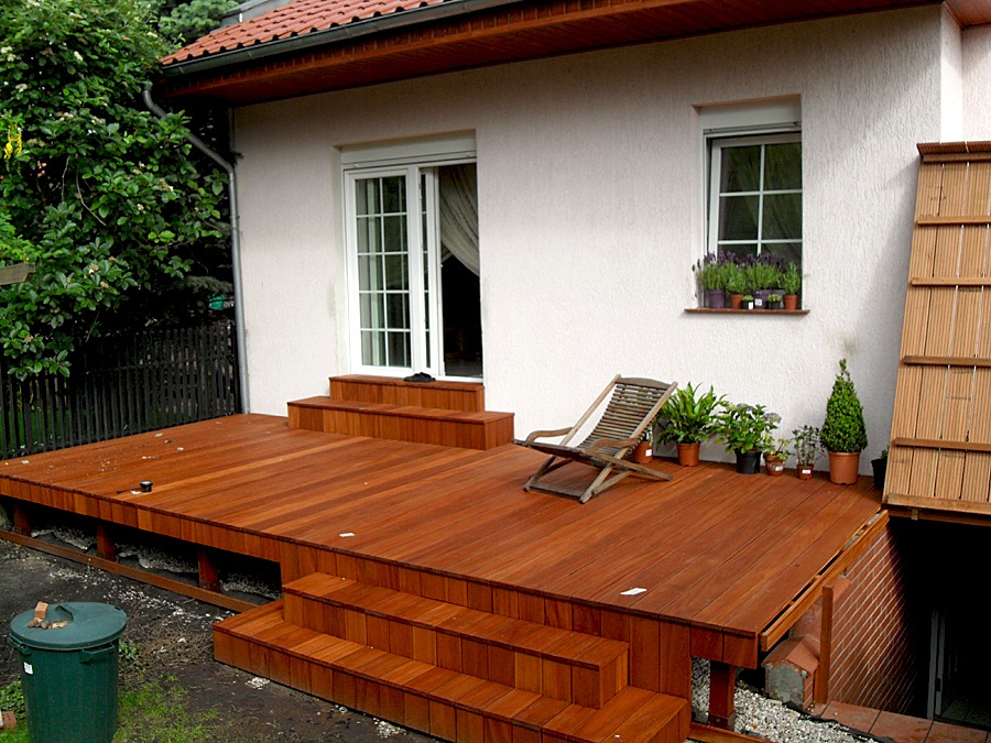 Gotowy taras z drewna egzotycznego z niskimi schodami, z drewnianym leżakiem i doniczkami z roślinami na tle białej ściany domu z oknem i kwiatami.