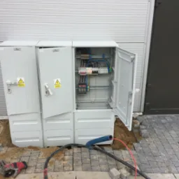 Otwarta skrzynka elektryczna z widocznymi bezpiecznikami i przewodami, umieszczona w wykopie obok kostki brukowej, z roboczą rękawicą obok kabli na ziemi.