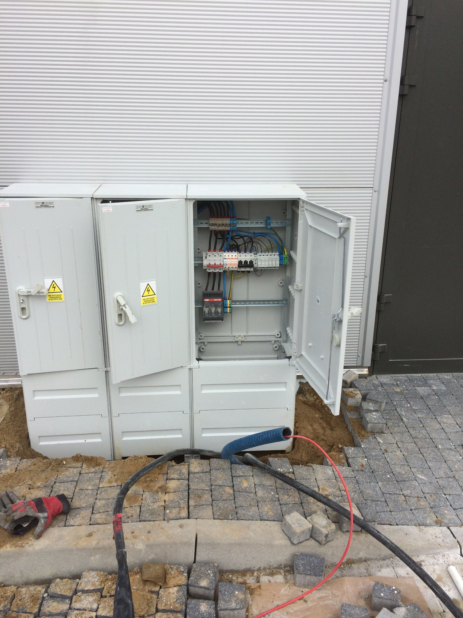 Otwarta skrzynka elektryczna z widocznymi bezpiecznikami i przewodami, umieszczona w wykopie obok kostki brukowej, z roboczą rękawicą obok kabli na ziemi.