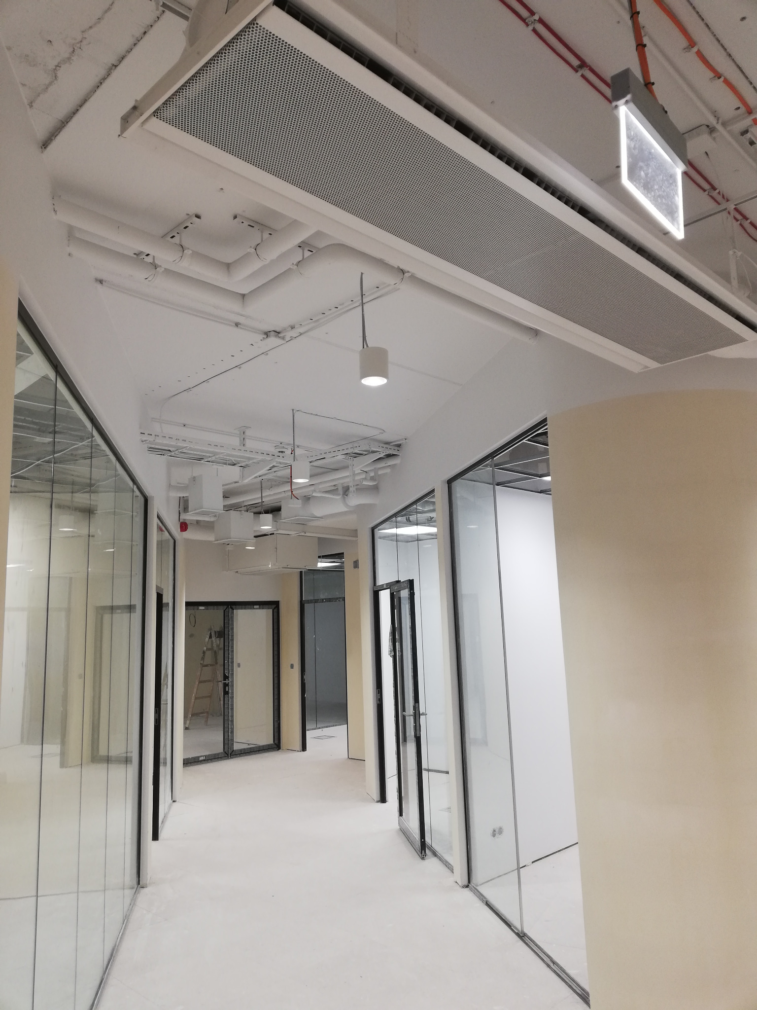 Korytarz biurowy z przeszklonymi ścianami działowymi i widocznymi instalacjami pod sufitem, w tym rurami, przewodami i kratkami wentylacyjnymi.