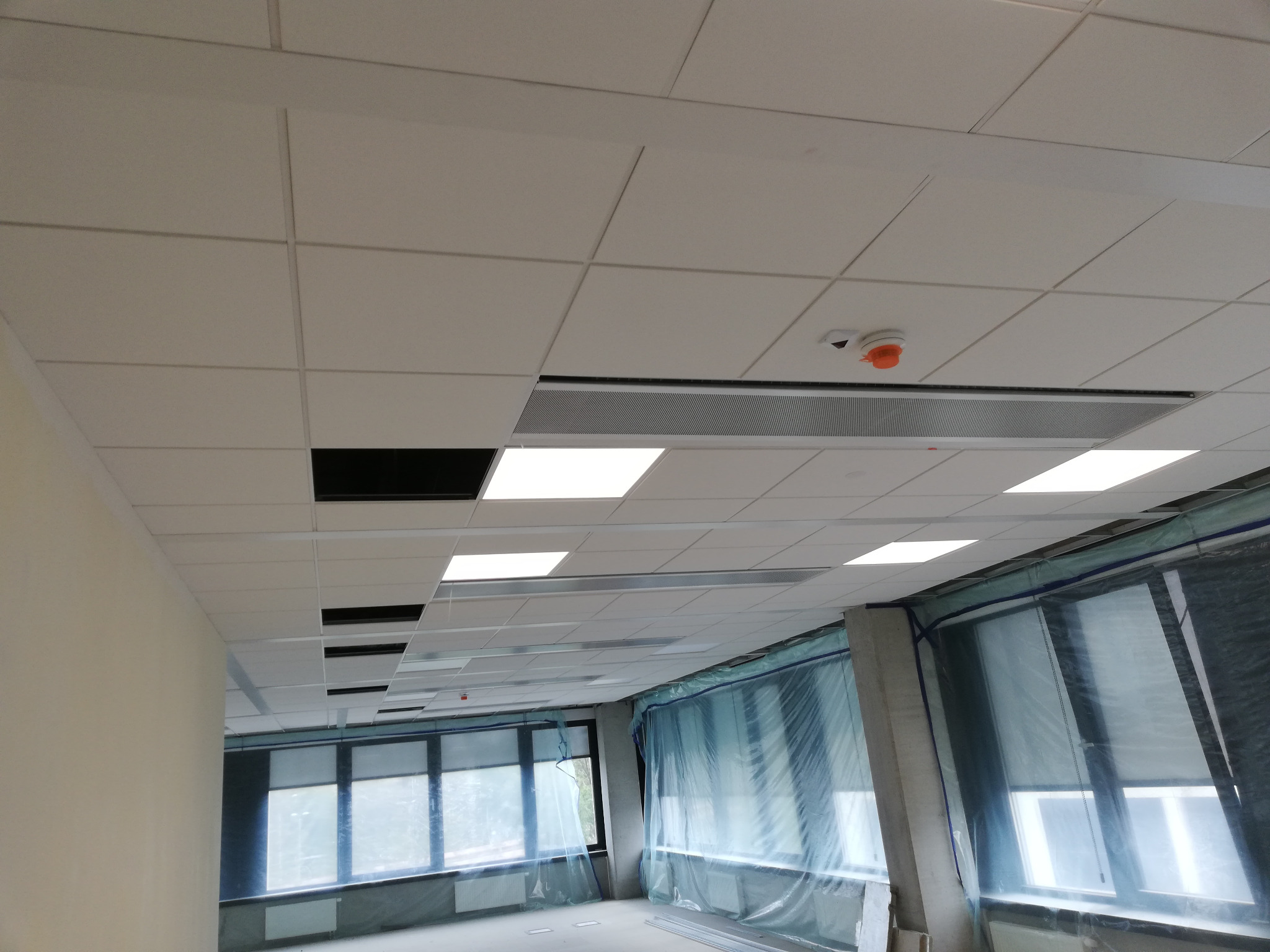 Sufit podwieszany z rastrem i oświetleniem LED w nowym biurze, widoczne okna zabezpieczone folią malarską.