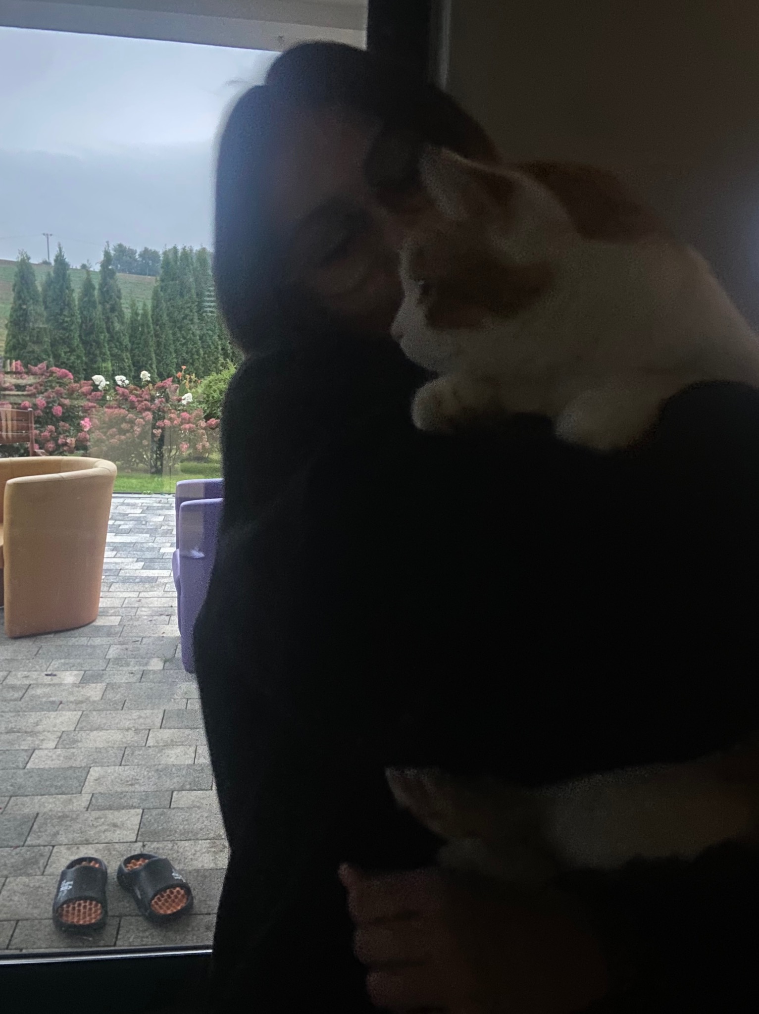 Kobieta w okularach przytula rudego kota na tle ogrodu z kwitnącymi różami, widoczne krzesła ogrodowe i klapki na podłodze.