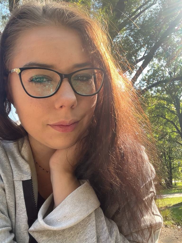 Portret kobiety w okularach na tle słonecznego parku, z refleksami na szkłach okularów.