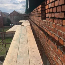 Murowana ściana domu w trakcie budowy, widoczna z poziomu rusztowania, z fragmentami niezamkniętych otworów okiennych i widocznymi śladami zaprawy.