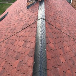 PVK Group Sp z o p - Monter w trakcie montażu kalenicy na dachu pokrytym czerwoną dachówką bitumiczną, widoczny komin z cegły i fragment budynku w tle.