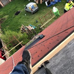 PVK Group Sp z o p - Praca na dachu z czerwonymi gontami bitumicznymi w trakcie wymiany, widoczny fragment stopy w sportowym bucie, młotek dekarski, narzędzia i samochód firmy w tle.