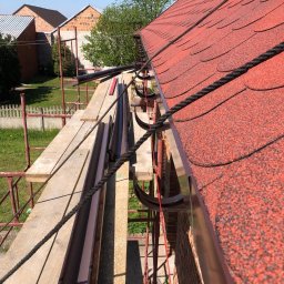 PVK Group Sp z o p - Czerwony dach z gontu bitumicznego, zamontowany nowy system rynnowy w kolorze brązowym, widok z rusztowania, w tle budynki mieszkalne.