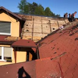 Demontaż starego dachu z czerwonej dachówki bitumicznej na domu jednorodzinnym, widoczne deski poszycia, dwóch dekarzy pracuje na górze, cień osoby robiącej zdjęcie na pierwszym planie.