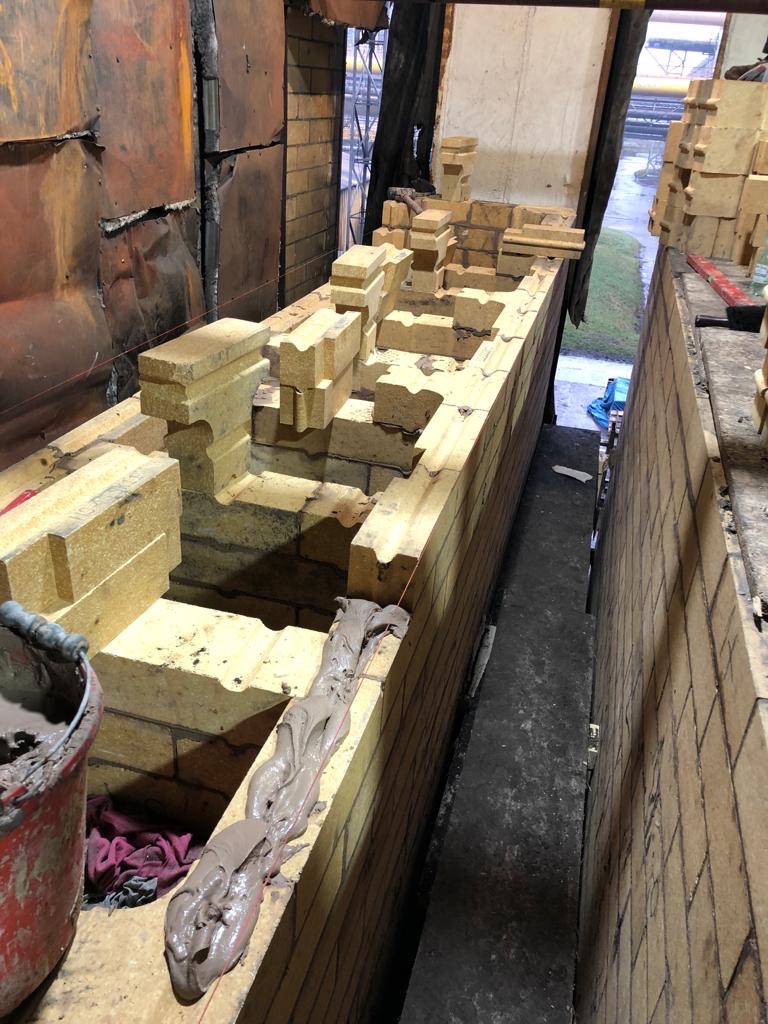 Wnętrze pieca przemysłowego w trakcie budowy z cegieł szamotowych, widoczna zaprawa murarska i wiadro z zaprawą, w tle konstrukcja stalowa i zieleń.