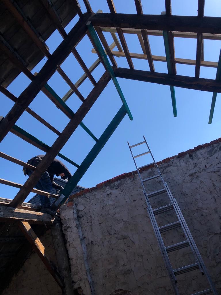 Pracownik w trakcie naprawy drewnianej konstrukcji dachu z widoczną drabiną opartą o mur budynku, na tle błękitnego nieba.