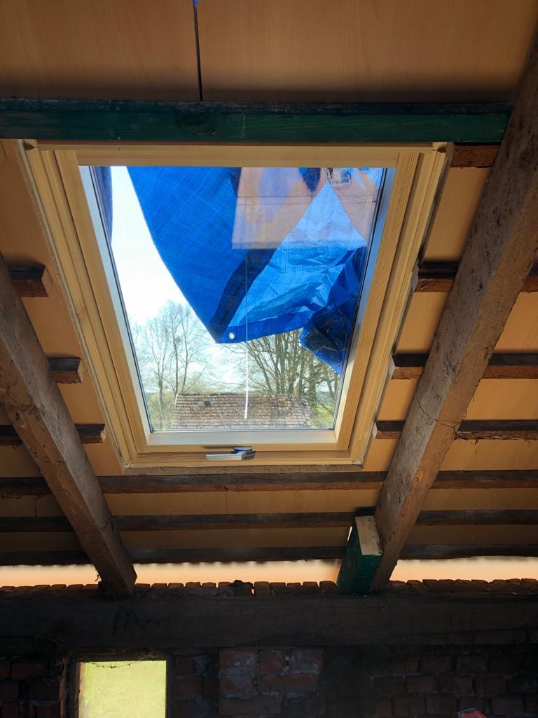 Wnętrze poddasza z drewnianą konstrukcją i nowym oknem dachowym, zabezpieczonym niebieską folią, widok na drzewa i dach budynku na zewnątrz.