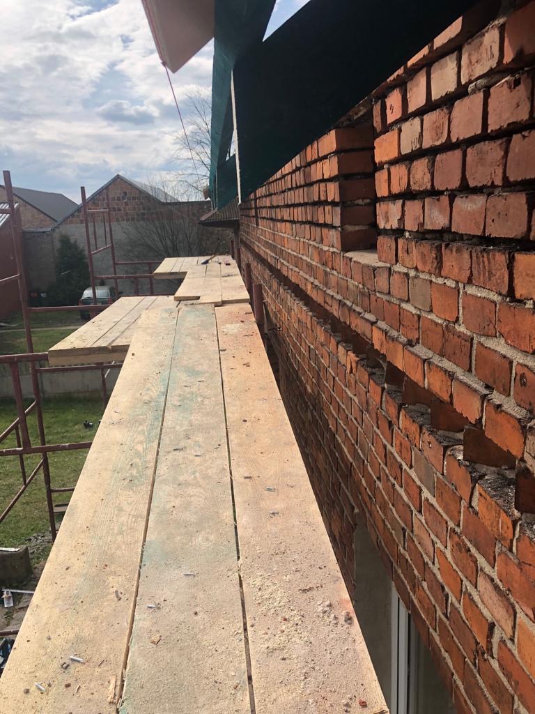 Murowana ściana domu w trakcie budowy, widoczna z poziomu rusztowania, z fragmentami niezamkniętych otworów okiennych i widocznymi śladami zaprawy.