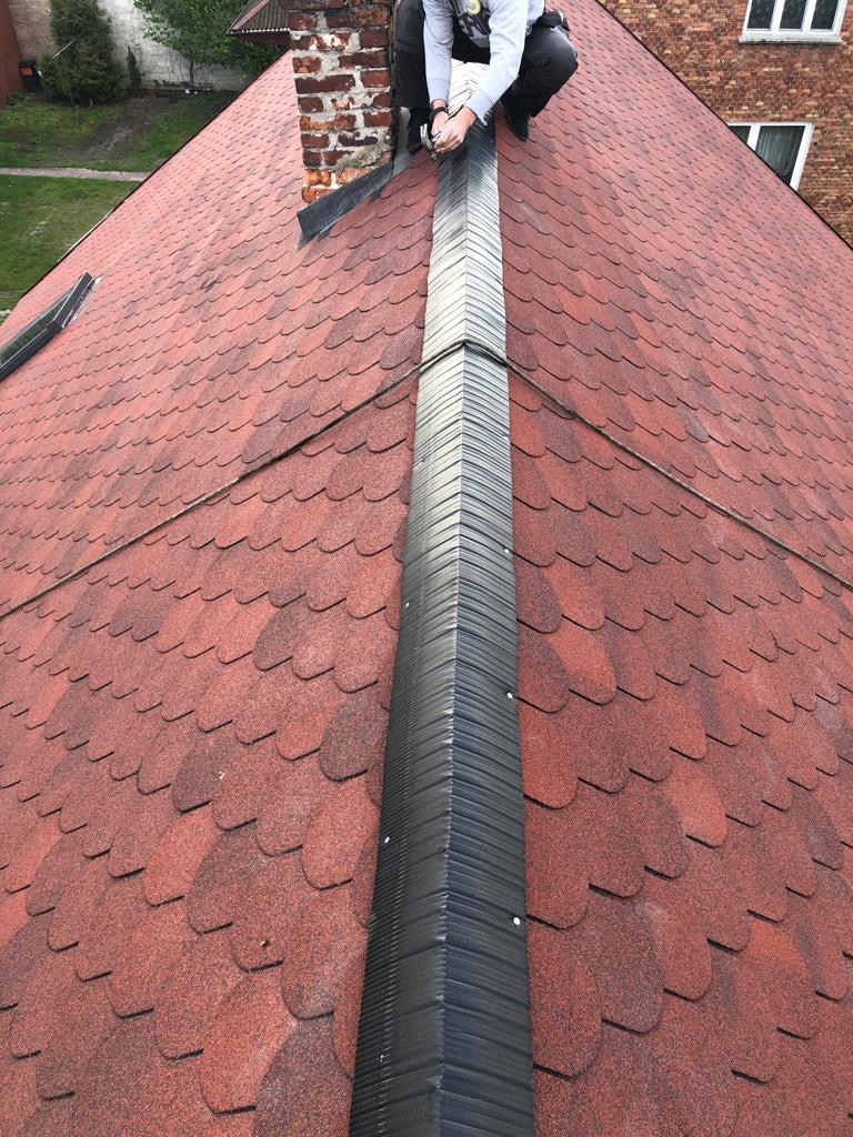Monter w trakcie montażu kalenicy na dachu pokrytym czerwoną dachówką bitumiczną, widoczny komin z cegły i fragment budynku w tle.