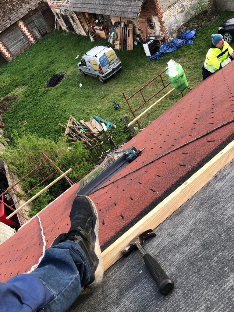 Praca na dachu z czerwonymi gontami bitumicznymi w trakcie wymiany, widoczny fragment stopy w sportowym bucie, młotek dekarski, narzędzia i samochód firmy w tle.