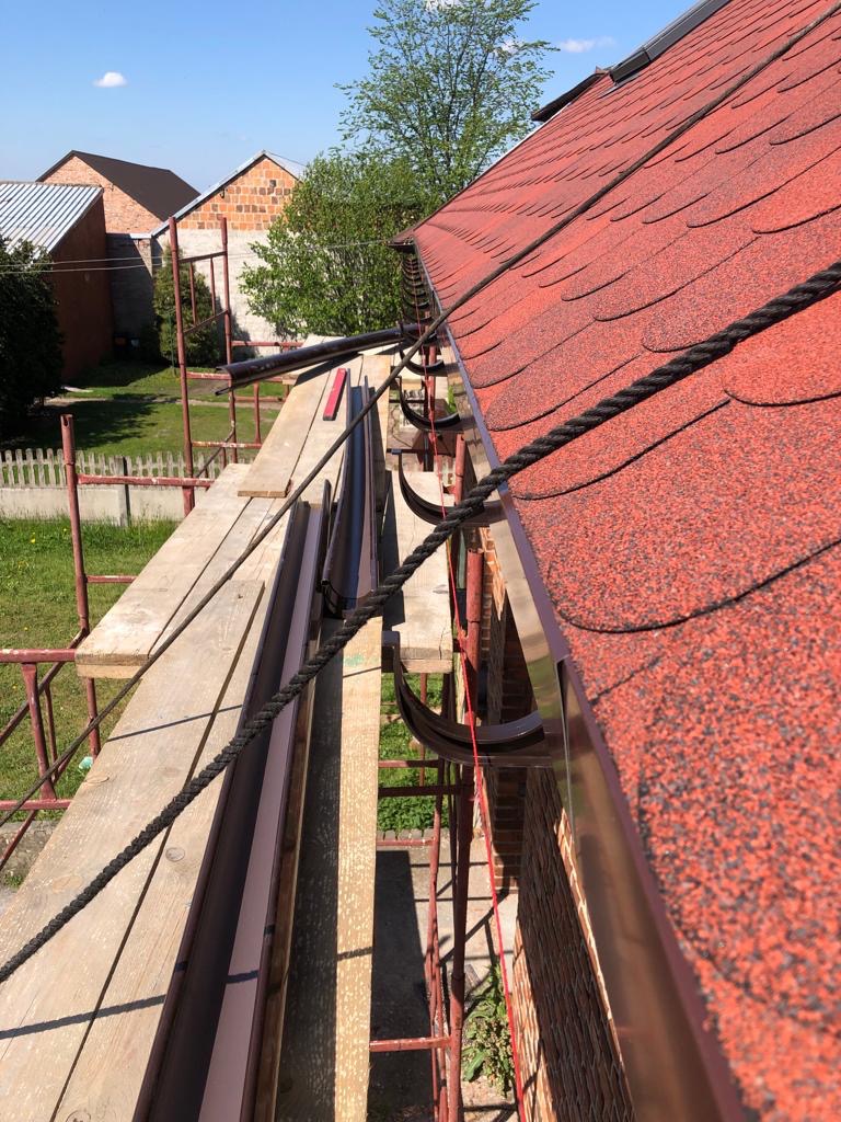 Czerwony dach z gontu bitumicznego, zamontowany nowy system rynnowy w kolorze brązowym, widok z rusztowania, w tle budynki mieszkalne.
