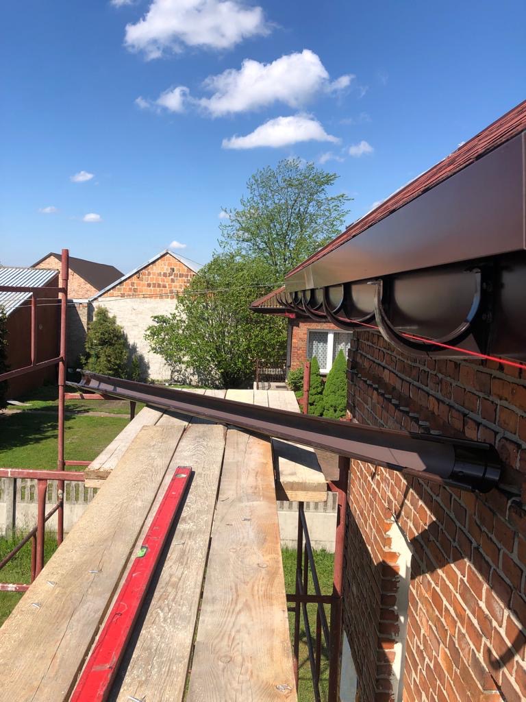 Montaż brązowych rynien na ceglanym domu w Sosnowcu, widok z rusztowania z poziomnicą.