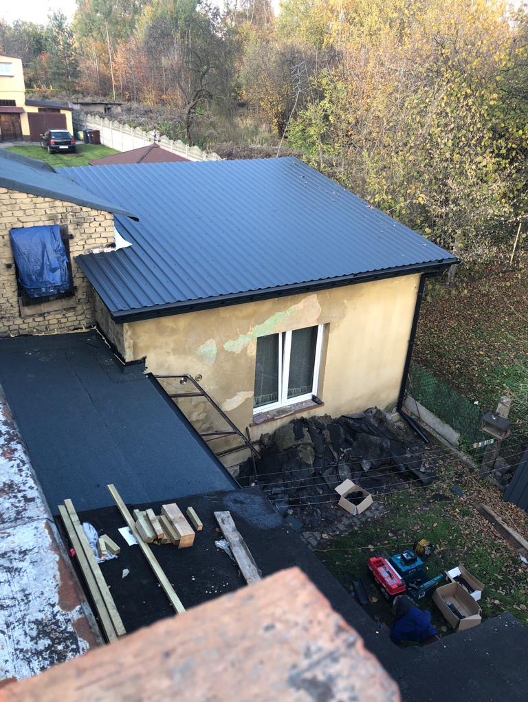 Widok z góry na budynek z nowym, ciemnoniebieskim dachem z blachy trapezowej; obok leżą materiały budowlane i narzędzia, a ściana budynku jest w trakcie remontu.