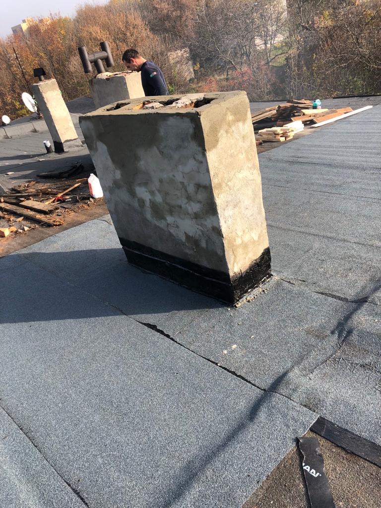 Prace dekarskie: Wykończony komin na dachu pokrytym papą, widoczny dekarz przy kolejnym kominie, pozostałości materiałów budowlanych na dachu.