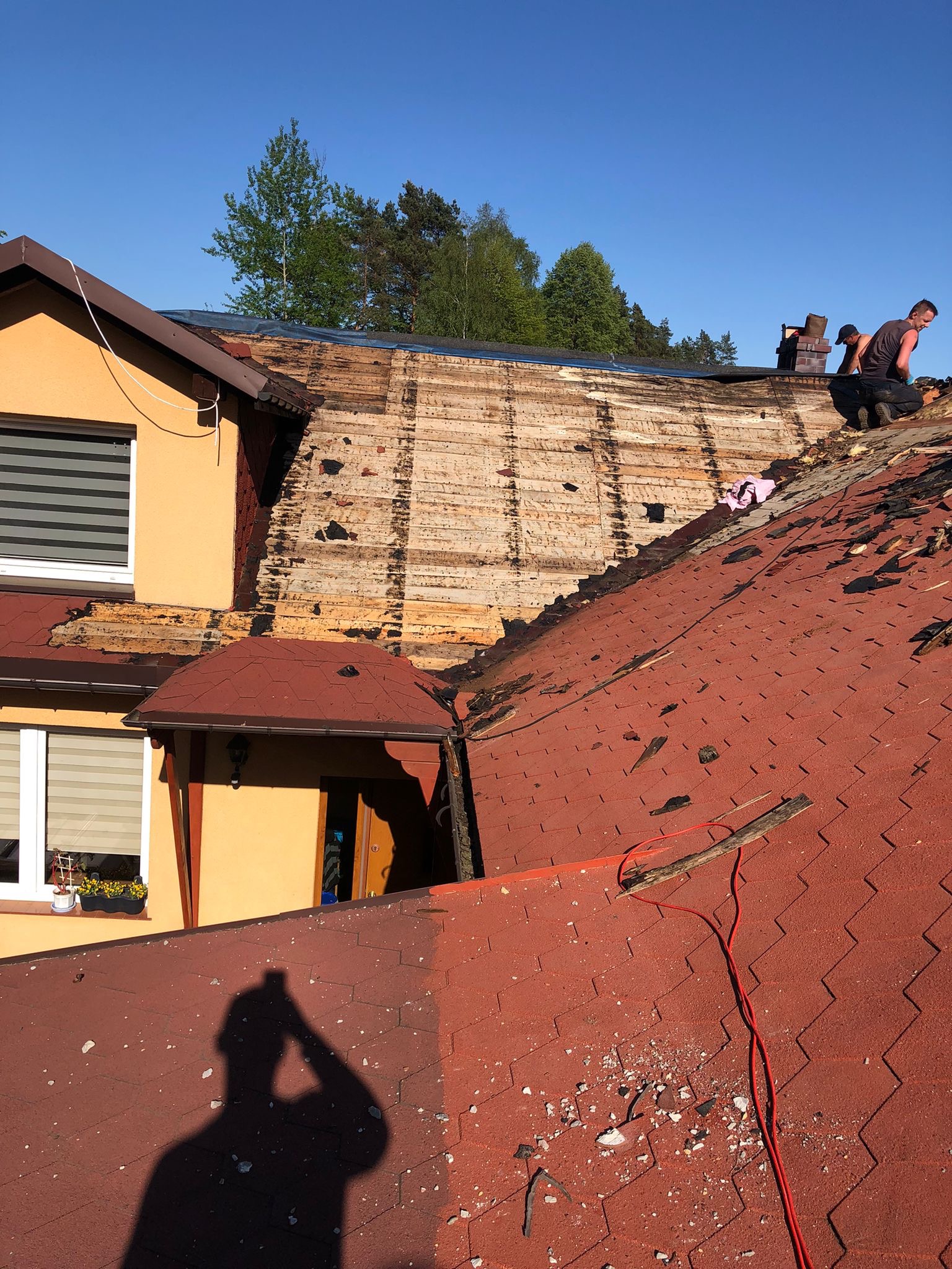 Demontaż starego dachu z czerwonej dachówki bitumicznej na domu jednorodzinnym, widoczne deski poszycia, dwóch dekarzy pracuje na górze, cień osoby robiącej zdjęcie na pierwszym planie.