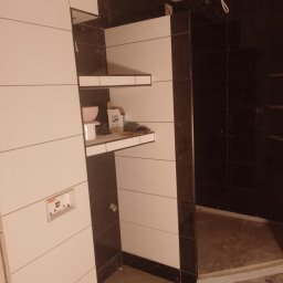 Pavel Druzhynskyi - Biało-czarna łazienka w trakcie wykańczania, widoczne płytki na ścianach, półki z materiałami budowlanymi, podłoga z poziomującymi krzyżykami do płytek.