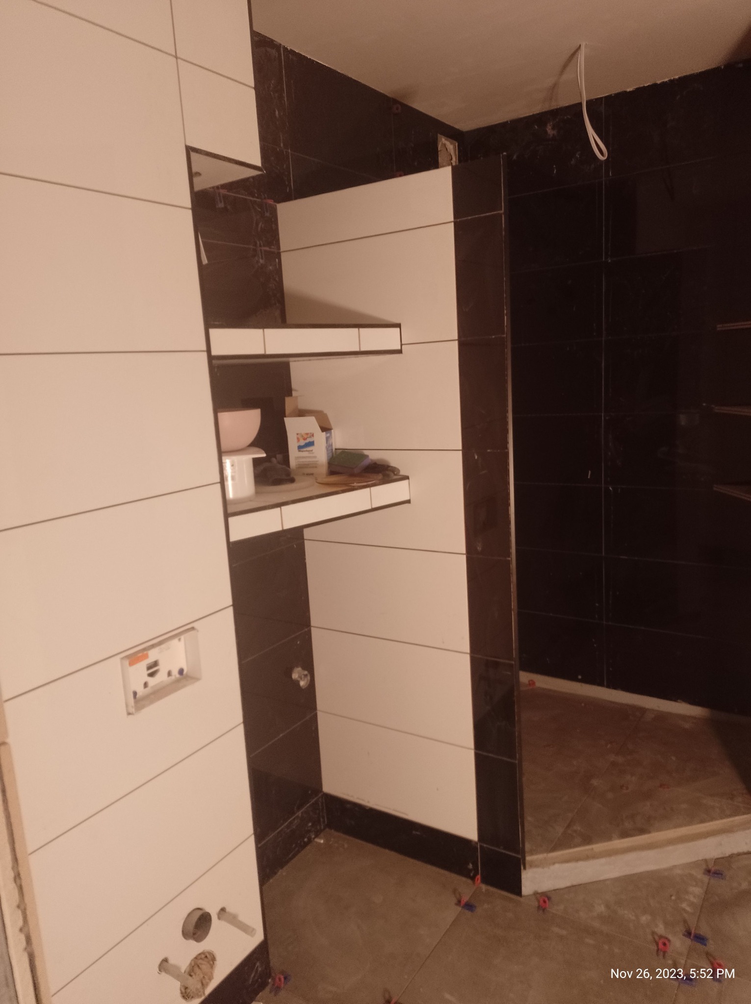 Biało-czarna łazienka w trakcie wykańczania, widoczne płytki na ścianach, półki z materiałami budowlanymi, podłoga z poziomującymi krzyżykami do płytek.